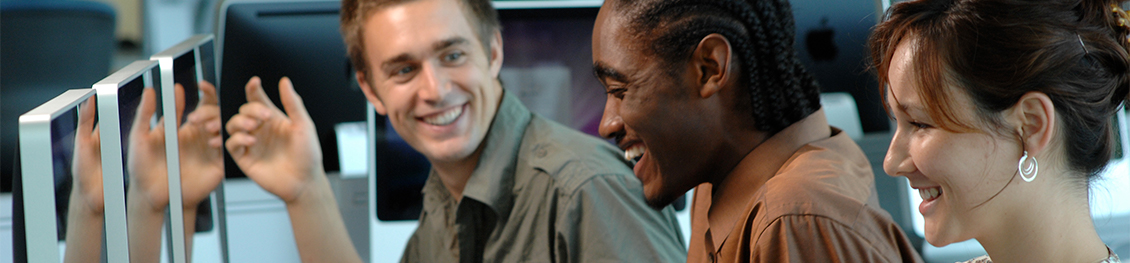 A white male student, a black male student, and a white female student laughing while working on computers